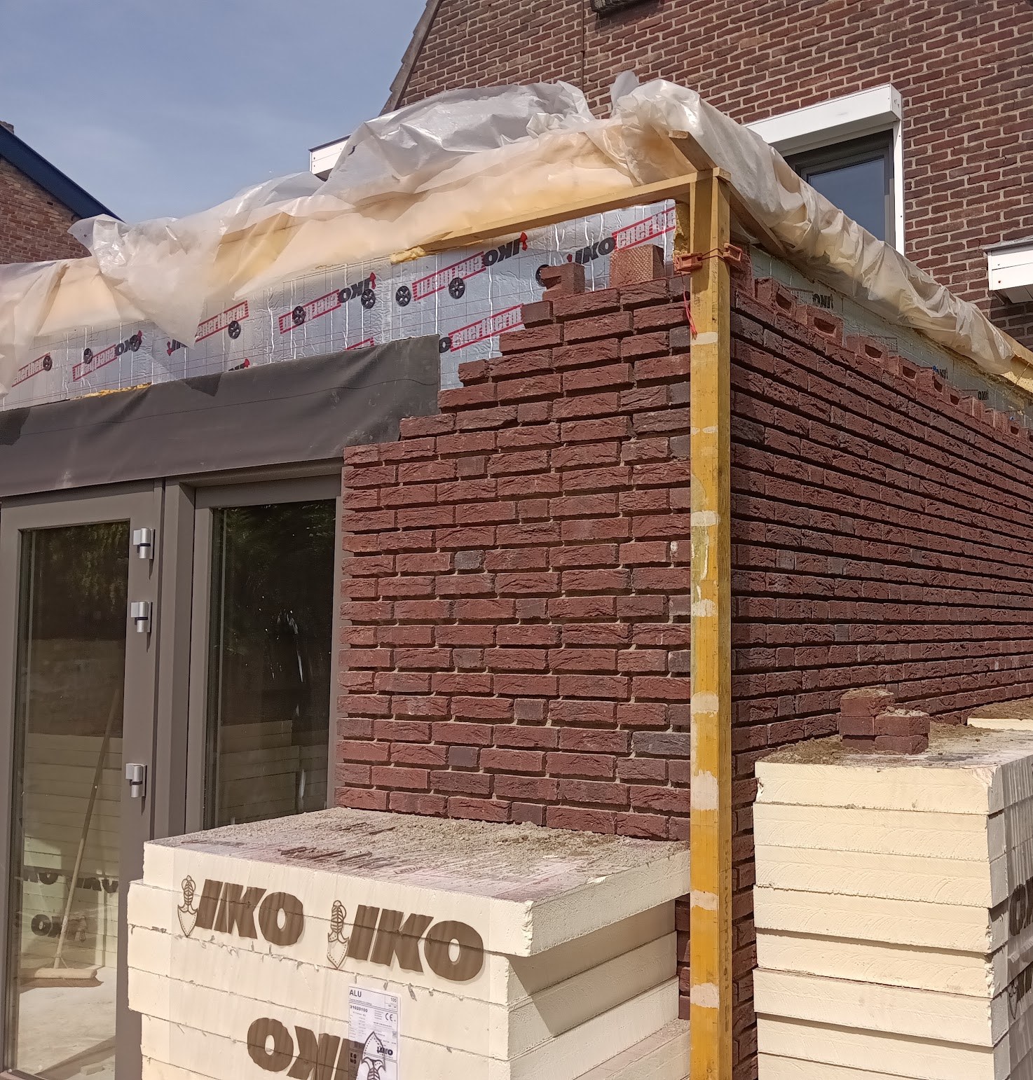 Gevel van een aanbouw in aanbouw: rode baksteen gaat omhoog, isolatieplaten liggen klaar