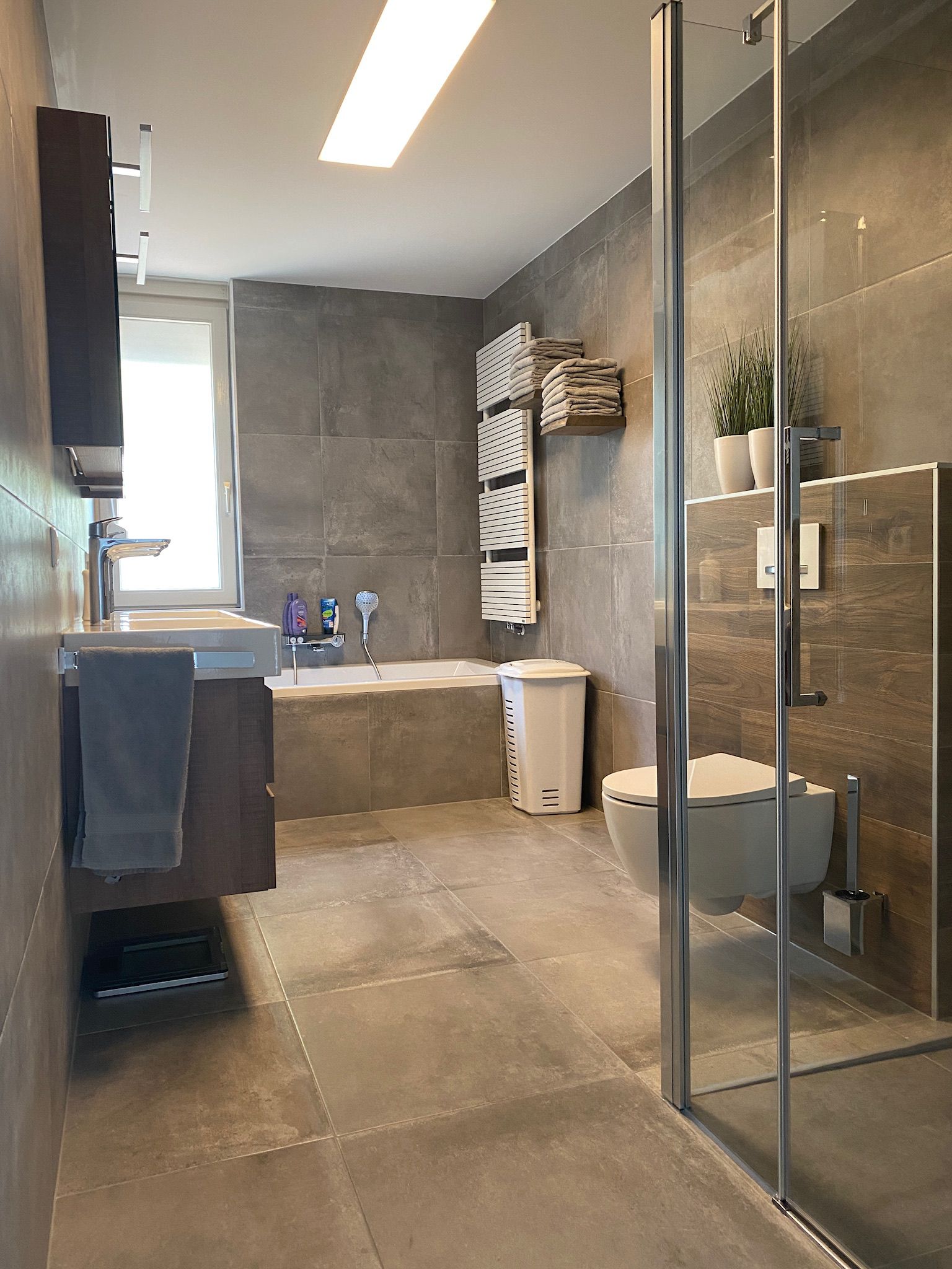Badkamer met grijze tegels, ligbad, glazen douchewand en handdoekradiator